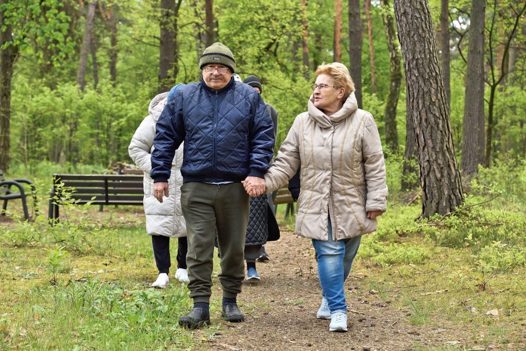 Grupa seniorów spaceruje leśną ścieżką podczas zajęć rekreacyjnych organizowanych przez ośrodek rehabilitacyjny. Na pierwszym planie starsza para trzyma się za ręce i rozmawia, a w tle widać ławki oraz zielony las