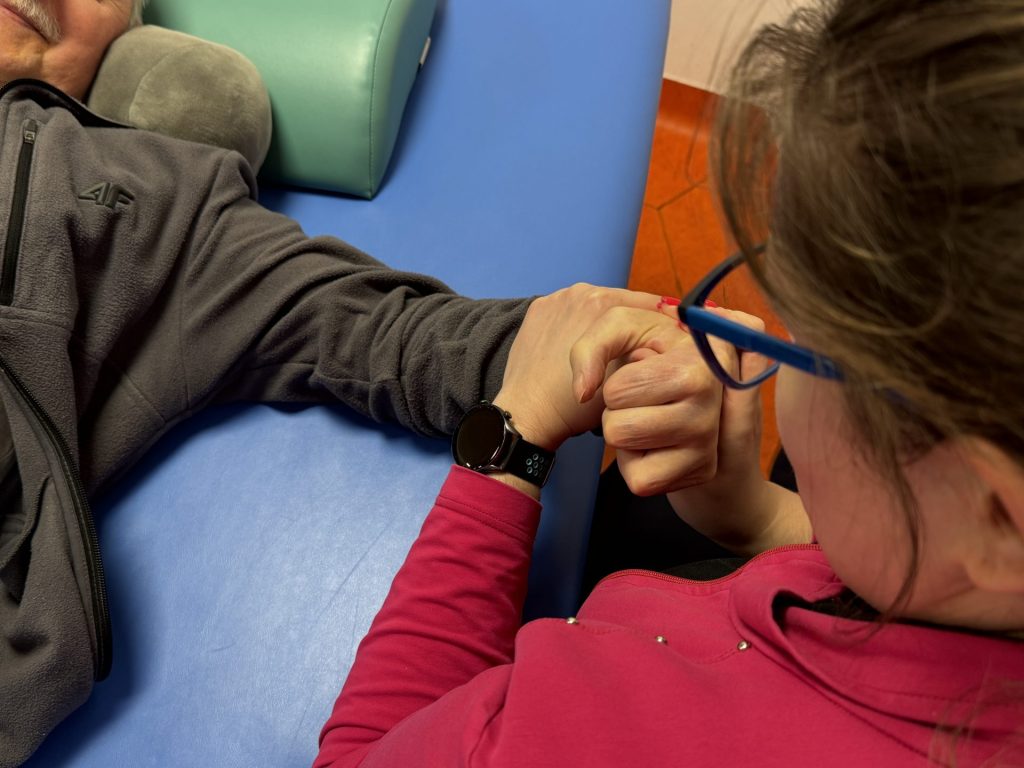 Ćwiczenia ręki podczas rehabilitacji po udarze. Pacjent leży na stole terapeutycznym, a fizjoterapeutka prowadzi terapię dłoni, wspierając ruch i chwyt w obrębie nadgarstka oraz palców. Zbliżenie pokazuje pracę manualną nad poprawą sprawności kończyny górnej