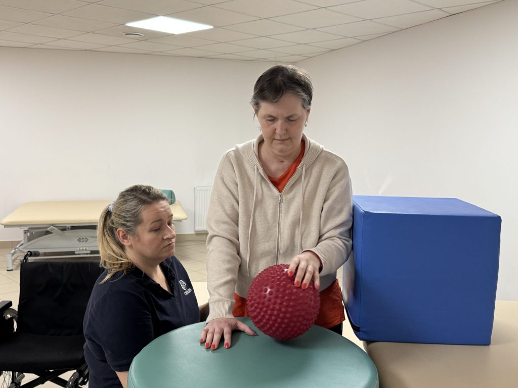 Ćwiczenia ręki podczas rehabilitacji po udarze. Pacjentka wykonuje zadanie z piłką sensoryczną przy stole terapeutycznym, a fizjoterapeutka obserwuje i wspiera prawidłowe ustawienie dłoni oraz kontrolę ruchu w sali rehabilitacyjnej