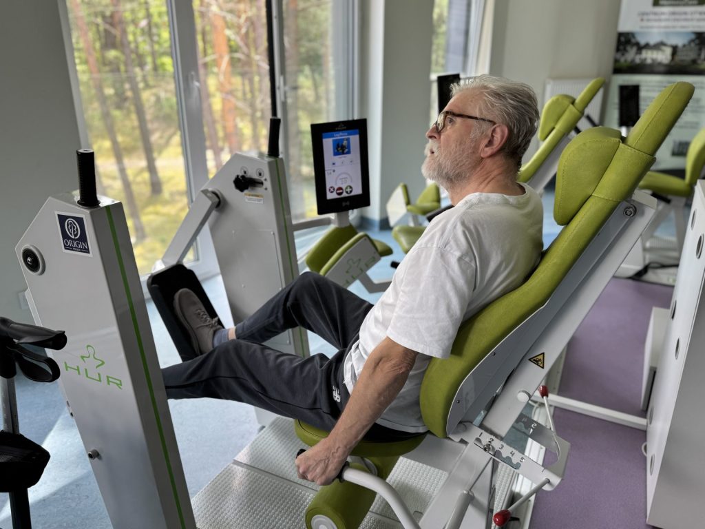 Starszy mężczyzna ćwiczy na urządzeniu do treningu nóg w nowoczesnej sali rehabilitacyjnej. Siedzi na specjalistycznym sprzęcie przy dużym oknie z widokiem na las, wykonując ćwiczenia wzmacniające nogi.