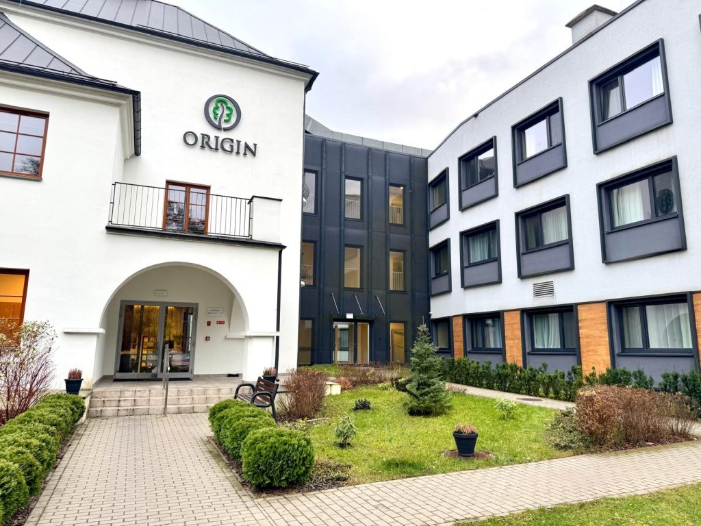 Budynek ośrodka rehabilitacyjnego Origin. Nowoczesny obiekt z jasną elewacją i widocznym logo nad wejściem, otoczony zielenią oraz zadbanym dziedzińcem prowadzącym do głównego wejścia placówki