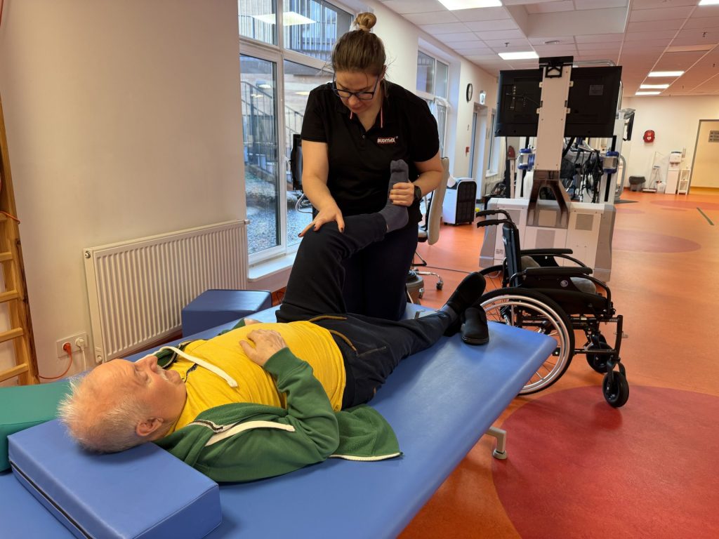 Fizjoterapia podczas rehabilitacji po udarze w sali ćwiczeń. Starszy mężczyzna leży na stole terapeutycznym, a fizjoterapeutka wykonuje ćwiczenia bierne nogi, wspierając zgięcie w stawie kolanowym. W tle widoczny jest wózek inwalidzki oraz specjalistyczny sprzęt rehabilitacyjny