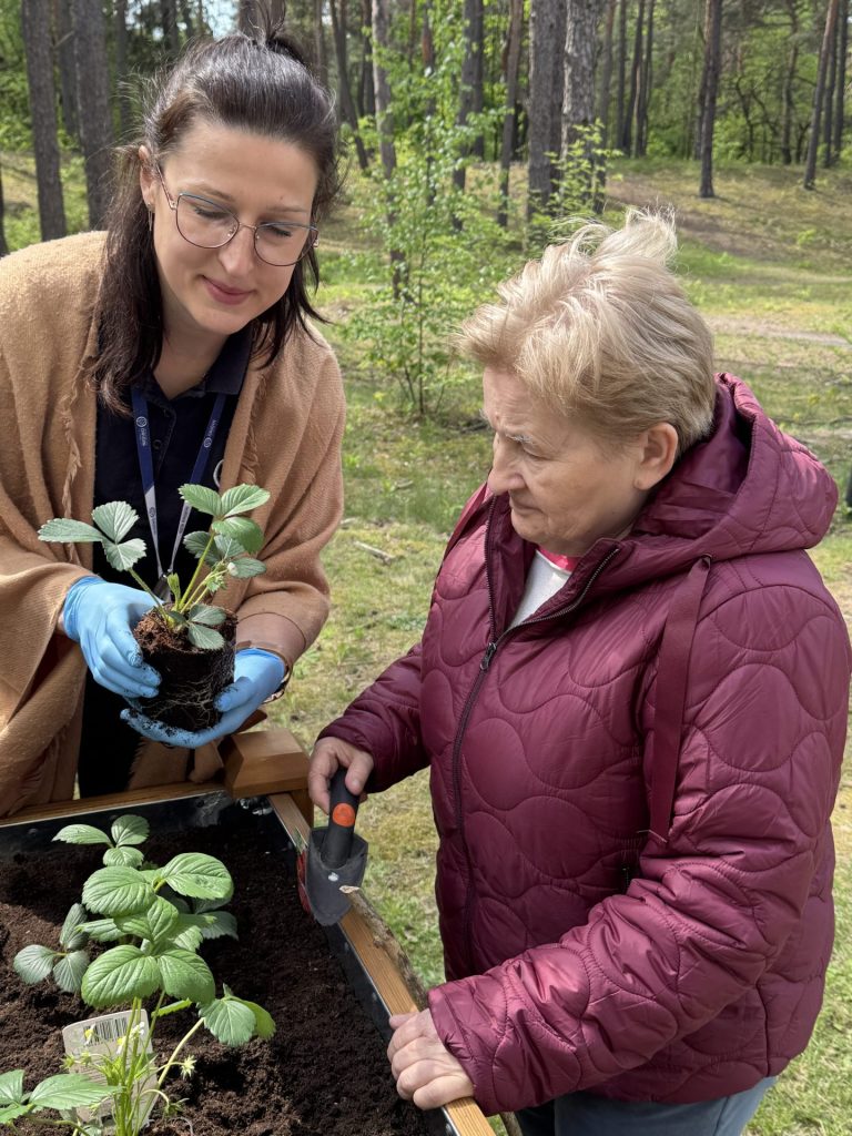 Terapeutka w rękawiczkach pokazuje starszej kobiecie sadzonkę truskawki podczas zajęć ogrodniczych na świeżym powietrzu przy ośrodku rehabilitacyjnym. Obok znajduje się podwyższona grządka z młodymi roślinami, a w tle widać las.