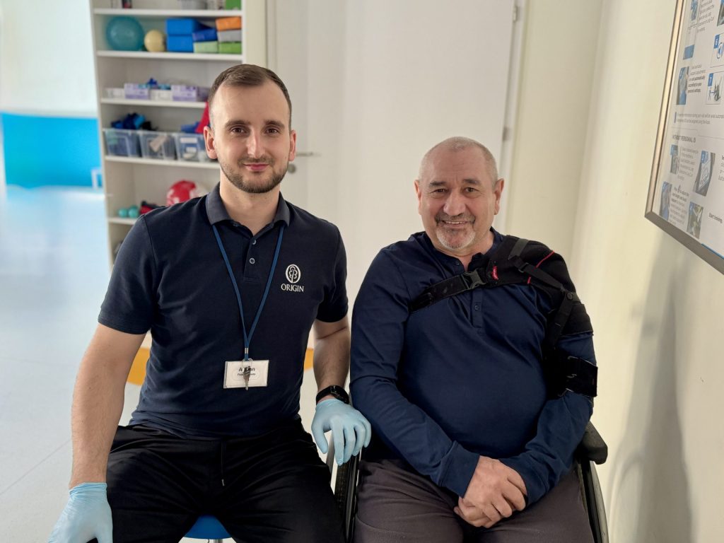Pacjent po udarze podczas rehabilitacji w ośrodku terapeutycznym. Mężczyzna siedzi na wózku inwalidzkim z ortezą stabilizującą bark, obok niego znajduje się fizjoterapeuta w stroju medycznym. Zdjęcie wykonane w sali rehabilitacyjnej.