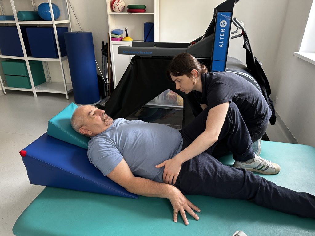 Ćwiczenia usprawniające podczas rehabilitacji po udarze. Pacjent leży na materacu z podparciem klinowym, a fizjoterapeutka prowadzi terapię kończyny dolnej, wspierając prawidłowe ustawienie biodra i kolana. W tle widoczny jest sprzęt do reedukcji chodu w sali rehabilitacyjnej