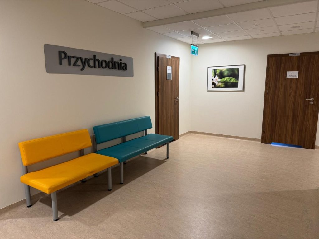 Poczekalnia w poradni logopedycznej i psychologicznej. Jasne wnętrze z kolorowymi ławkami dla pacjentów, drzwiami do gabinetów oraz oznaczeniem „Przychodnia” na ścianie.