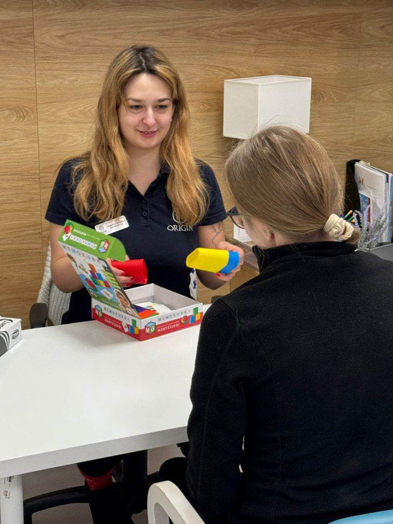 Zajęcia terapeutyczne podczas rehabilitacji po udarze. Specjalistka prowadzi ćwiczenia funkcji poznawczych przy biurku, prezentując kolorowe elementy edukacyjne pacjentce. Spotkanie odbywa się w gabinecie terapeutycznym, w spokojnej i wspierającej atmosferze