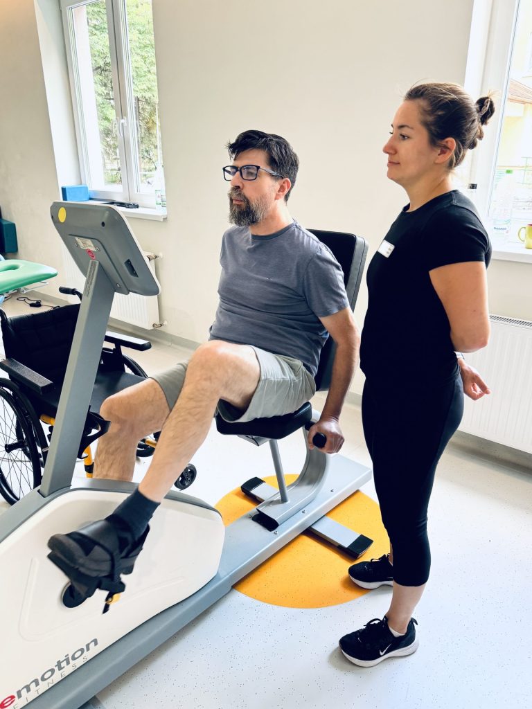 Trening na rowerze stacjonarnym podczas rehabilitacji po udarze. Młody pacjent wykonuje ćwiczenia wzmacniające kończyny dolne pod nadzorem fizjoterapeutki, która monitoruje prawidłową postawę i technikę ruchu w sali rehabilitacyjnej