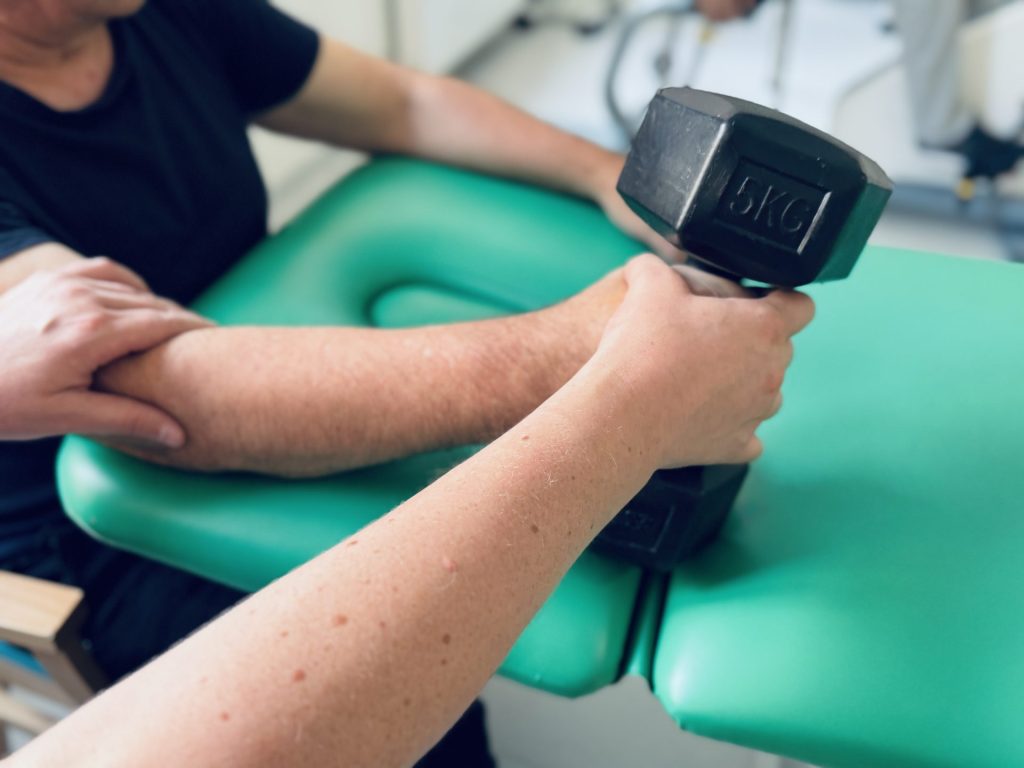 Wzmacnianie ręki podczas rehabilitacji po udarze. Zbliżenie na dłoń pacjenta wykonującego ćwiczenie z hantlem o wadze 5 kg, przy wsparciu terapeuty stabilizującego przedramię na stole rehabilitacyjnym.